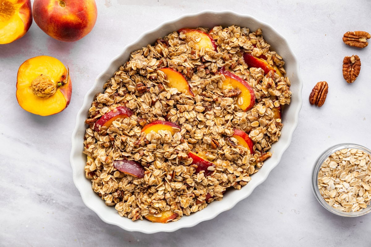 A peach crisp in a large oval baking dish with fresh peaches, pecans, and oats to the side.