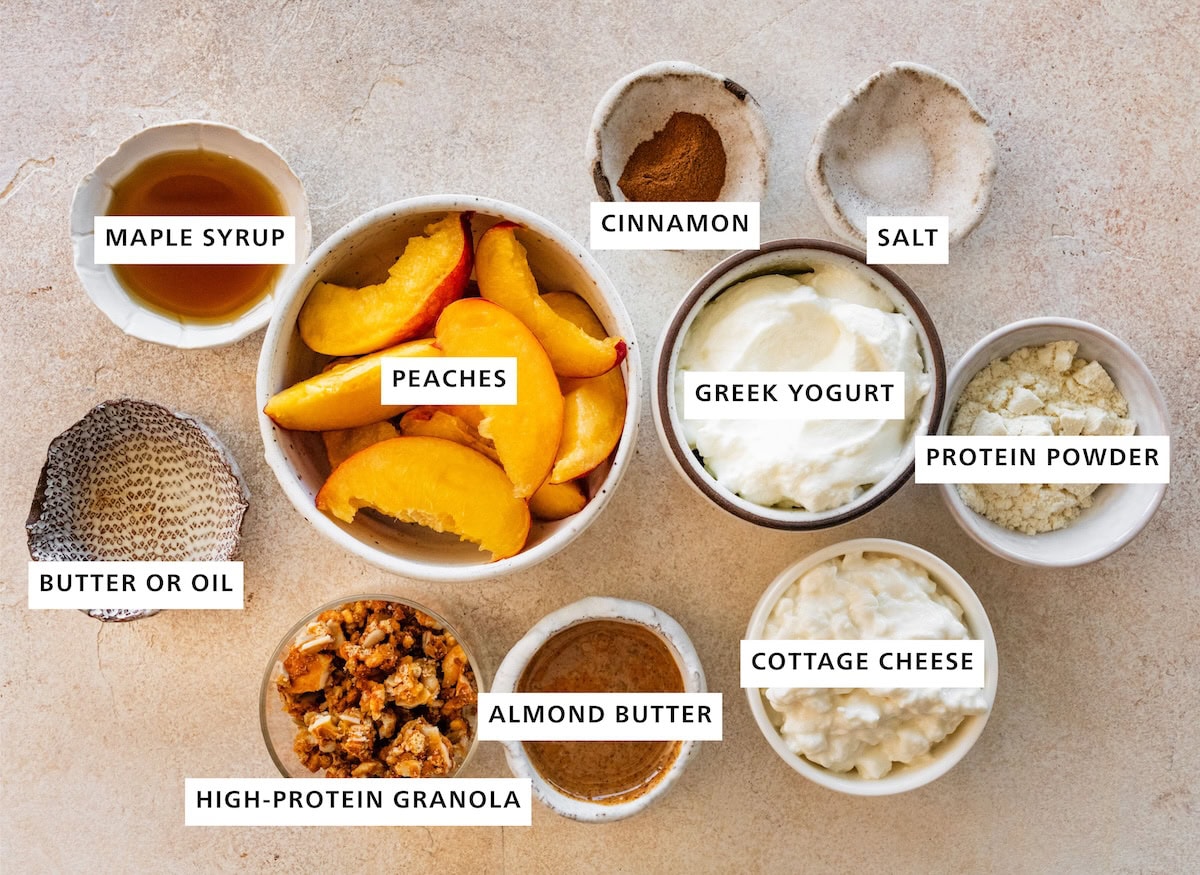 Ingredients measured out for a high-protein peach crisp yogurt bowl including: maple syrup, cinnamon, salt, peaches, greek yogurt, protein powder, cottage cheese, almond butter, high-protein granola, and butter/oil.
