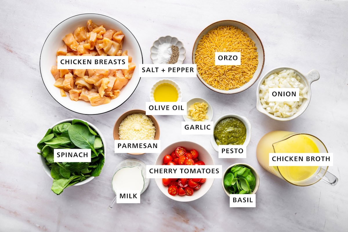Ingredients measured out for pesto chicken and orzo skillet: chicken breasts, salt, pepper, orzo, onion, chicken broth, basil, pesto, garlic, cherry tomatoes, milk, parmesan, olive oil, garlic, and spinach.