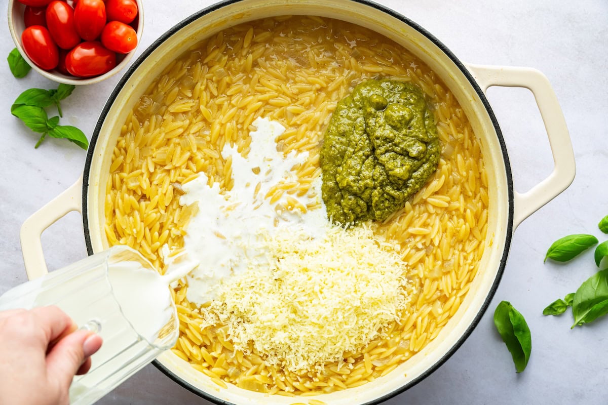 Milk is being poured into a large pot with cooked orzo, pesto, and parmesan cheese.