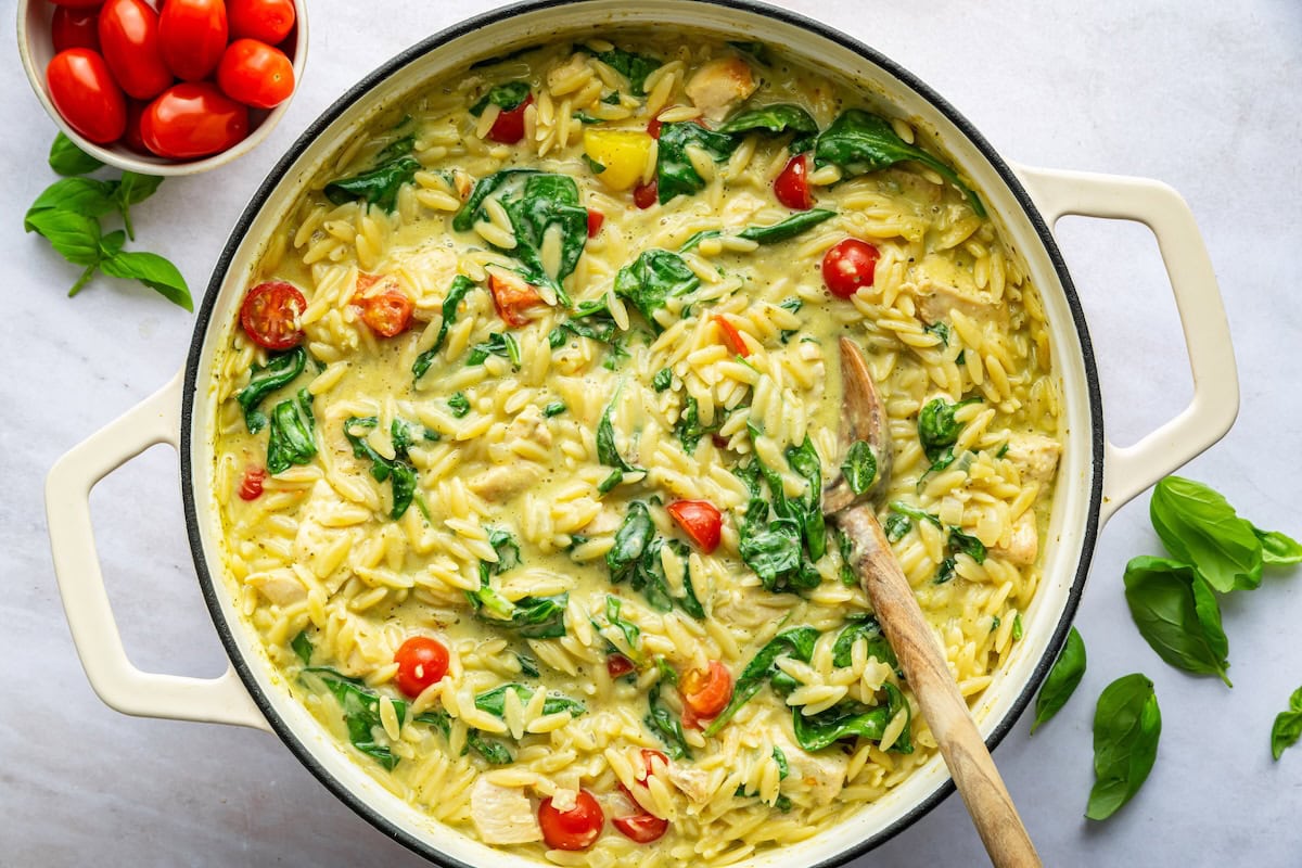 A large pot with pesto chicken, orzo, spinach, and cherry tomatoes with a wooden spoon.