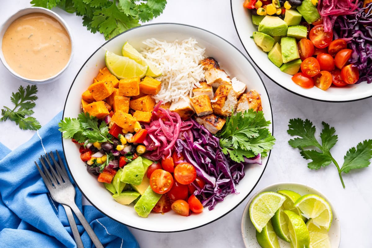 BBQ chicken bowls prepped and ready to serve with BBQ chicken, roasted sweet potatoes, cabbage, tomatoes, avocado, black bean and corn salad, rice and pickled red onions.