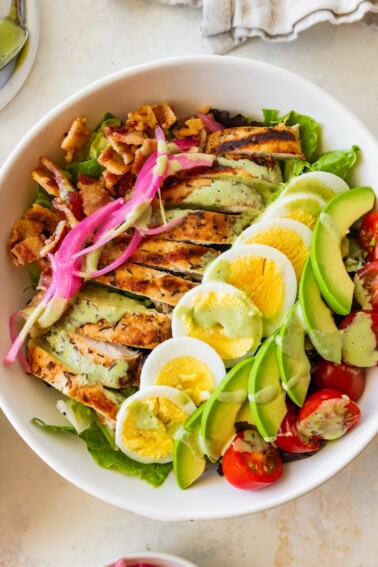 A chicken cobb salad with hard boiled egg, avocado, tomatoes, bacon, and onion in a bowl.