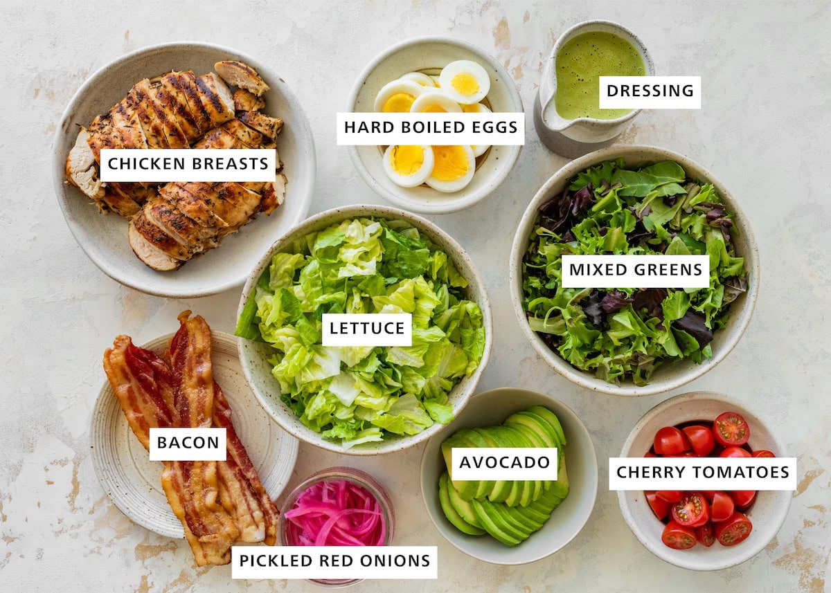 Ingredients measured out for green goddess cobb salad: chicken breasts, hard boiled eggs, dressing, lettuce, mixed greens, bacon, picked red onions, avocado, cherry tomatoes.