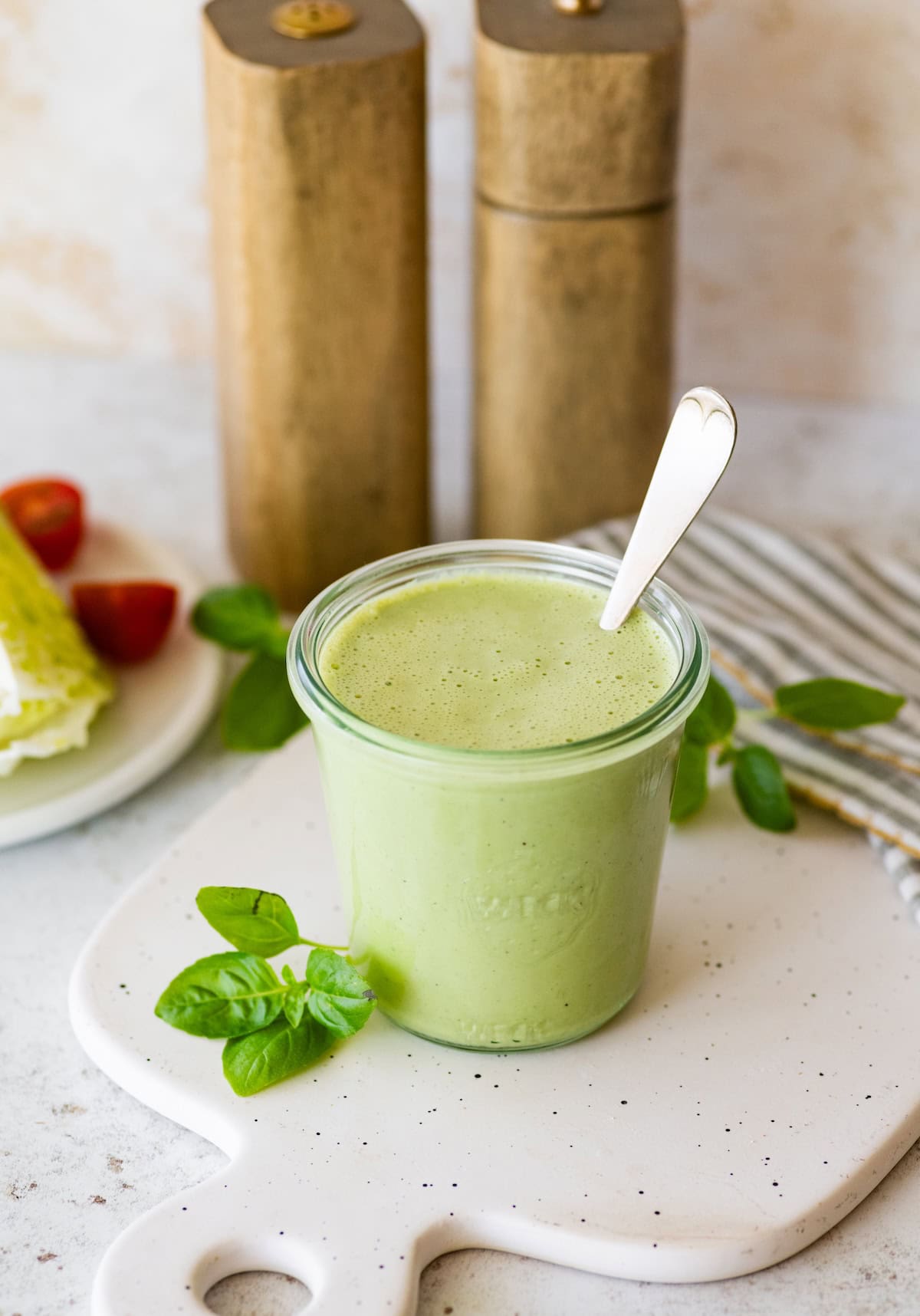 A green dressing in a glass jar with a metal spoon. The jar is on a cutting board with fresh basil leaves to the side.