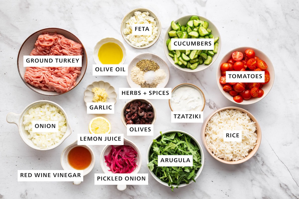 Ingredients measured out for ground turkey Greek bowls: ground turkey, olive oil, feta, cucumbers, tomatoes, rice, tzatziki, arugula, pickled onion, red wine vinegar, onion, lemon juice, olives, garlic, herbs, and spices.