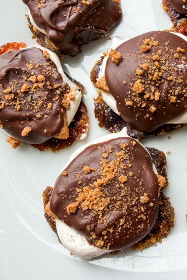 Smashed date s'mores on a white plate topped with melted chocolate and crushed graham crackers.